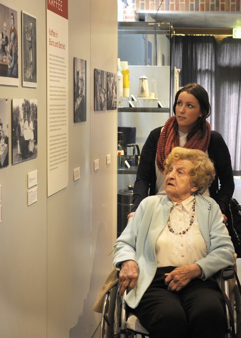 Eine junge Frau schiebt eine ältere Frau im Rollstuhl durch eine Ausstellung.