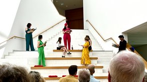 Auf einer Treppen stehen mehrere Menschen, die jeweils in einer Farbe gekleidet sind: rot, grün, gelb, blau, schwarz und weiß. Sie sprechen miteinander. Unten an der Treppe hochblickend sieht Publikum ihnen zu.